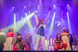 com um fundo lilás sob luzes dispersas, o Bando de Teatro Olodum se apresenta no palco. Eles olham para frente e fazes gesto de poder negro.