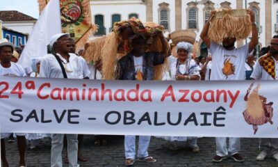 Fiéis de branco e com balaios de palha na cabeça atrás de uma faixa branca que diz "24ª caminhada azoany - salve obaluaiê"